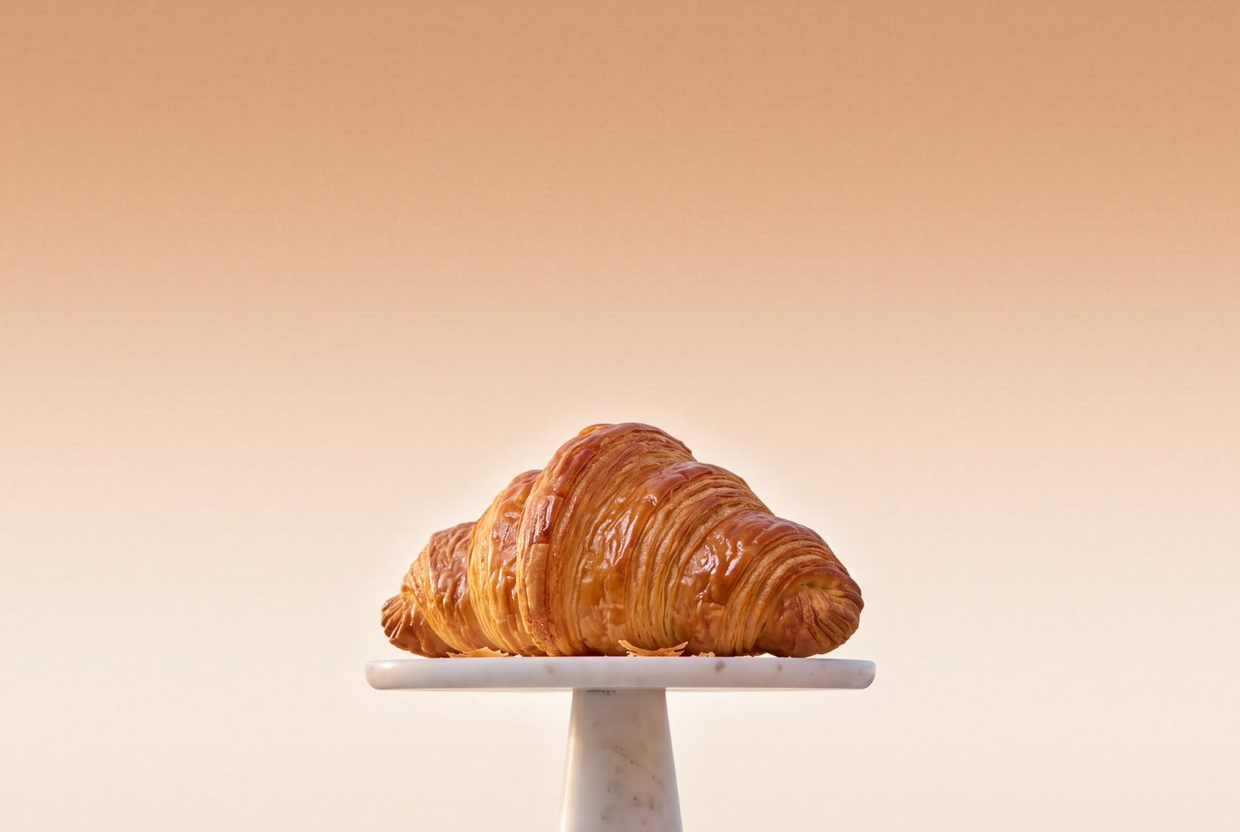A large golden all-butter croissant on a small marble stand, against a warm wheat-coloured sky.
